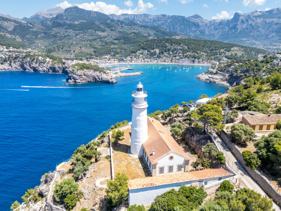 Aerial view of the Port de Sóller lighthouse overlooking turquoise waters, luxury yachts, and the Tramuntana mountains in Mallorca.