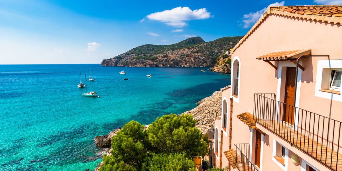 View of a Mediterranean coastline with turquoise waters, anchored boats, and pastel-colored houses overlooking the sea under a clear blue sky