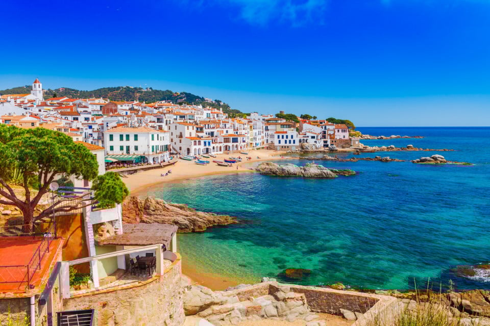 Panoramic view of a coastal village on the Costa Brava, with white houses overlooking the Mediterranean Sea, sandy beach and crystal-clear waters, one of the most attractive destinations to buy a second home in Spain.