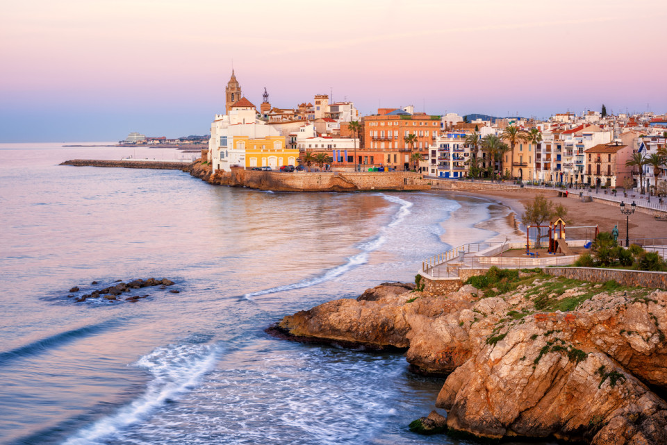 Sunset over Sitges shore, with calm sea reflecting golden and orange hues, and traditional Mediterranean houses lining the coast.
